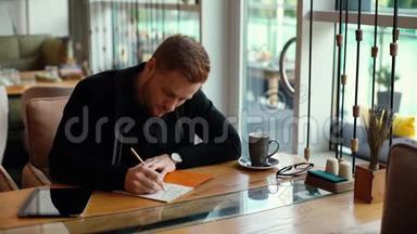 年轻的帅哥在笔记本上写字，在舒适的咖啡馆里写字